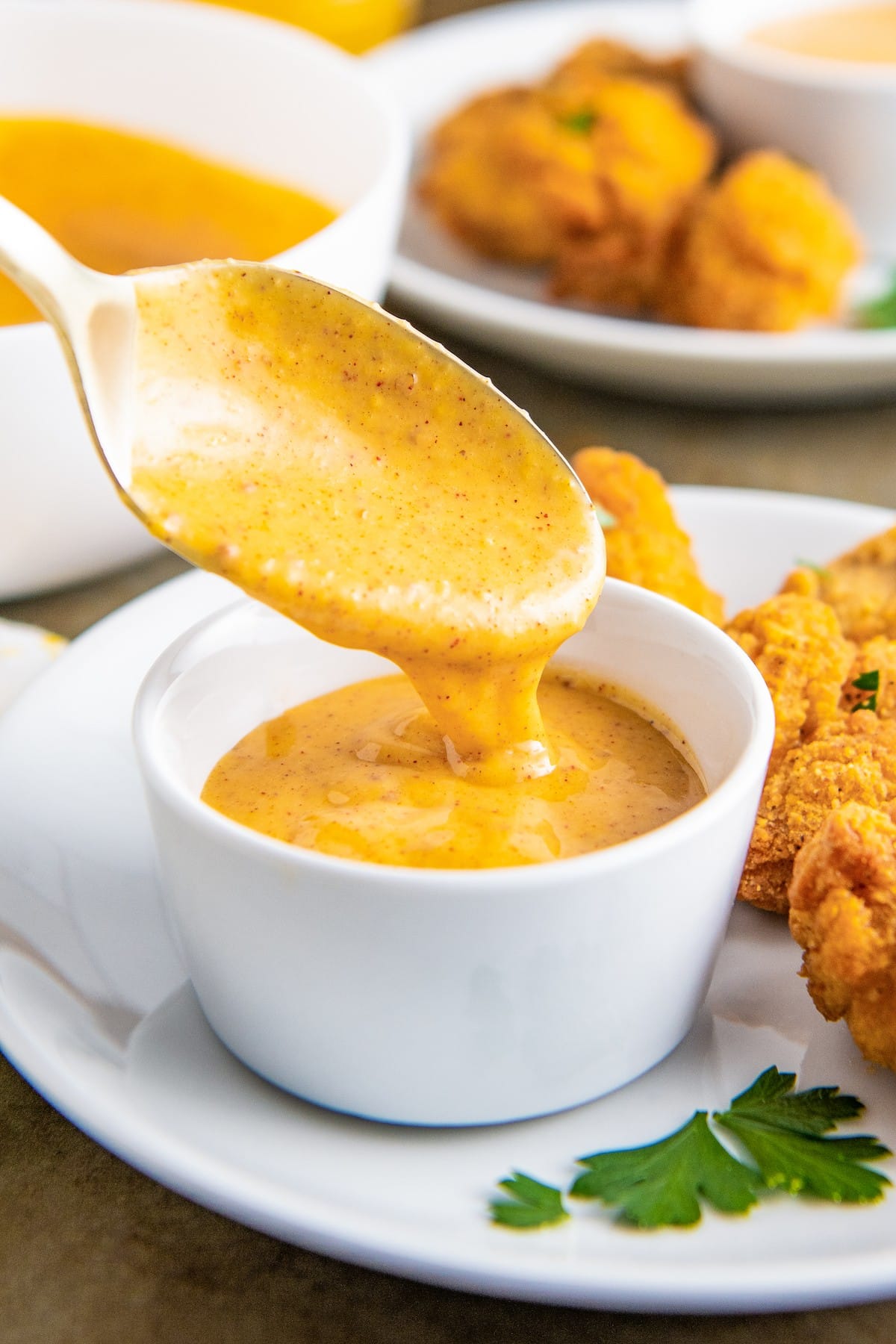 A spoon dripping honey mustard into a bowl of honey mustard, surrounded by nuggets