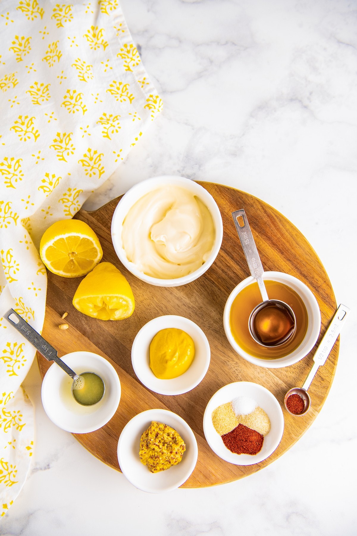 Overhead view of the ingredients needed for this honey mustard recipe, on a cutting board: a bowl of mayo, a lemon cut in half, a bowl of Dijon mustard, a bowl of yellow mustard, a bowl of lemon juice with a measuring spoon in it, a bowl of honey with a measuring spoon in it, a bowl of salt, onion powder, garlic powder, and paprika, and a measuring spoon with cayenne