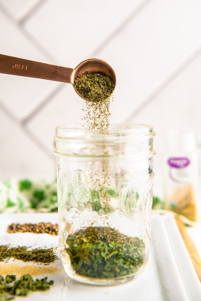 A spoon pouring herbs into a jar with herbs and spices