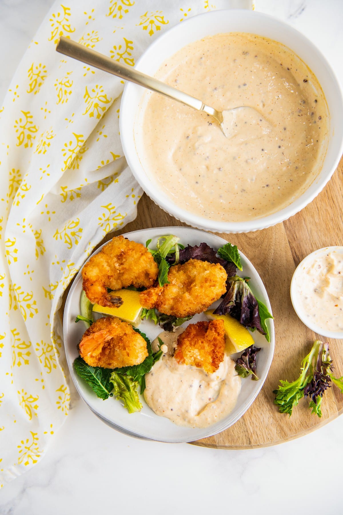 Overhead view of a bowl of remoulade sauce with a spoon in it, and a plate of fried shrimp, greens, lemons, and remoulade