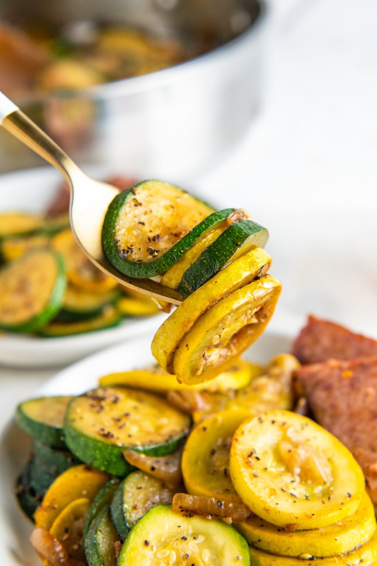 Sautéed zucchini and squash on a fork, above a plate full of squash, zucchini, and sausage