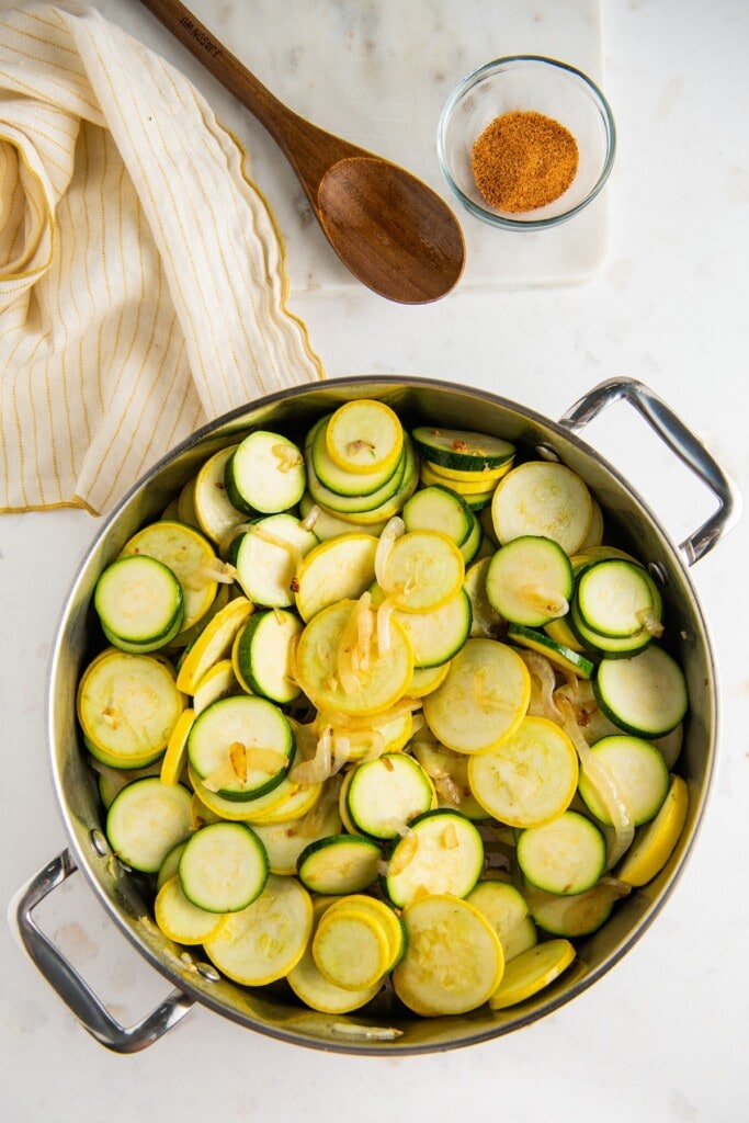Overhead view of a skillet filled with raw zucchini and squash rounds, next to a wooden spoon and a bowl of a spice rub