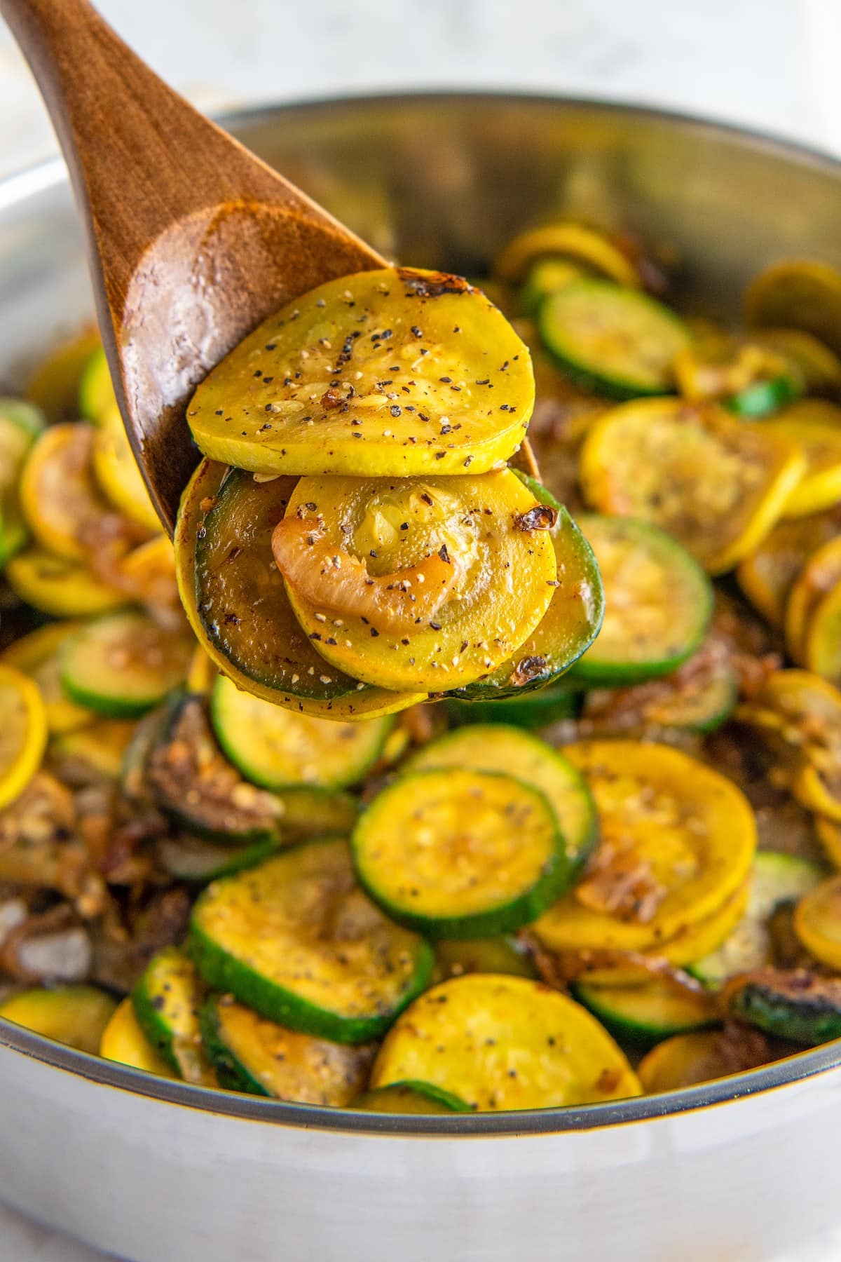 Close up of sautéed zucchini and squash on a wooden spoon, above a bowl full of zucchini and squash