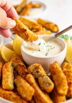 A hand dipping a zucchini fry into a bowl of ranch dressing surrounded by zucchini fries