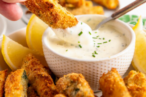 A hand dipping a zucchini fry into a bowl of ranch dressing surrounded by zucchini fries