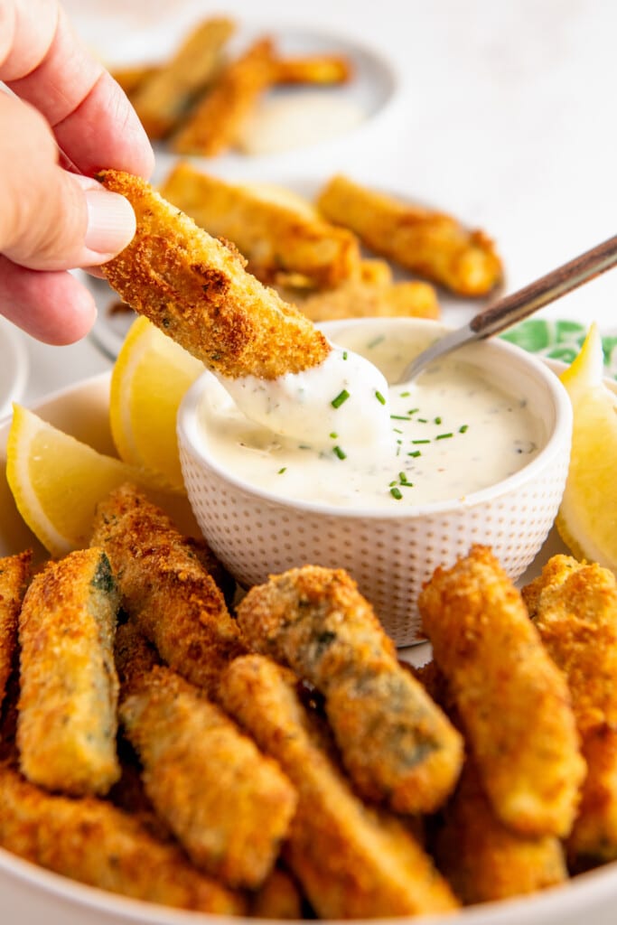 A hand dipping a zucchini fry into a bowl of ranch dressing surrounded by zucchini fries