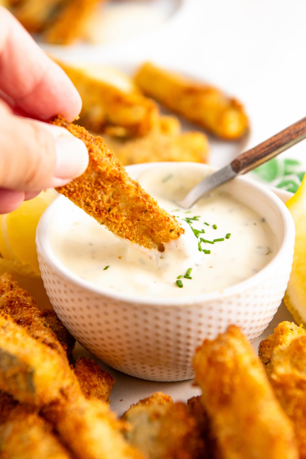 A hand dipping a zucchini fry into a bowl of ranch, surrounded by zucchini fries