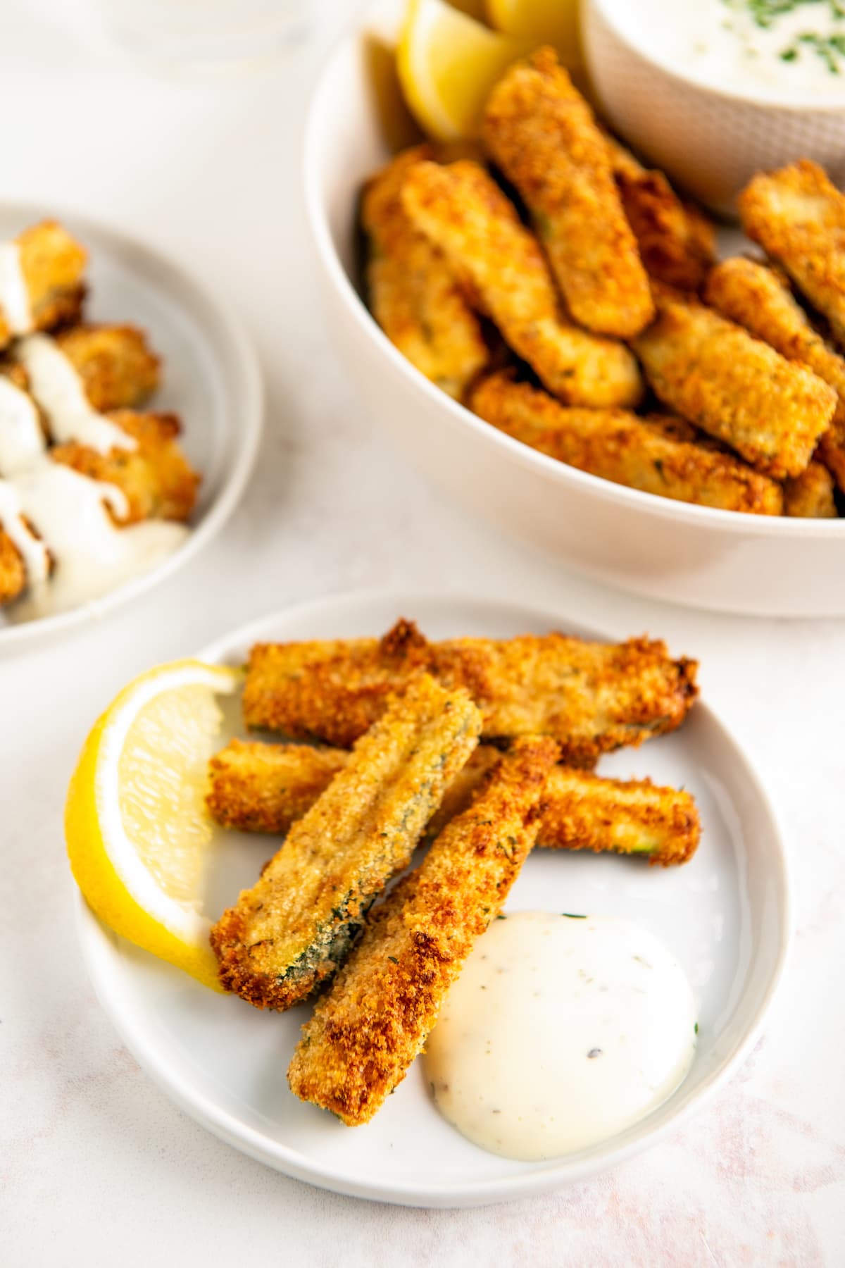 A plate of zucchini fries with a lemon wedge and a dollop of ranch, with a serving tray of zucchini fries in the background