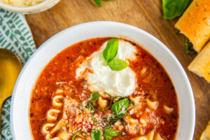 Overhead view of a bowl of lasagna soup topped with ricotta, parmesan, and basil, next to a bowl of parmesan cheese, a bowl of ricotta, and basil leaves