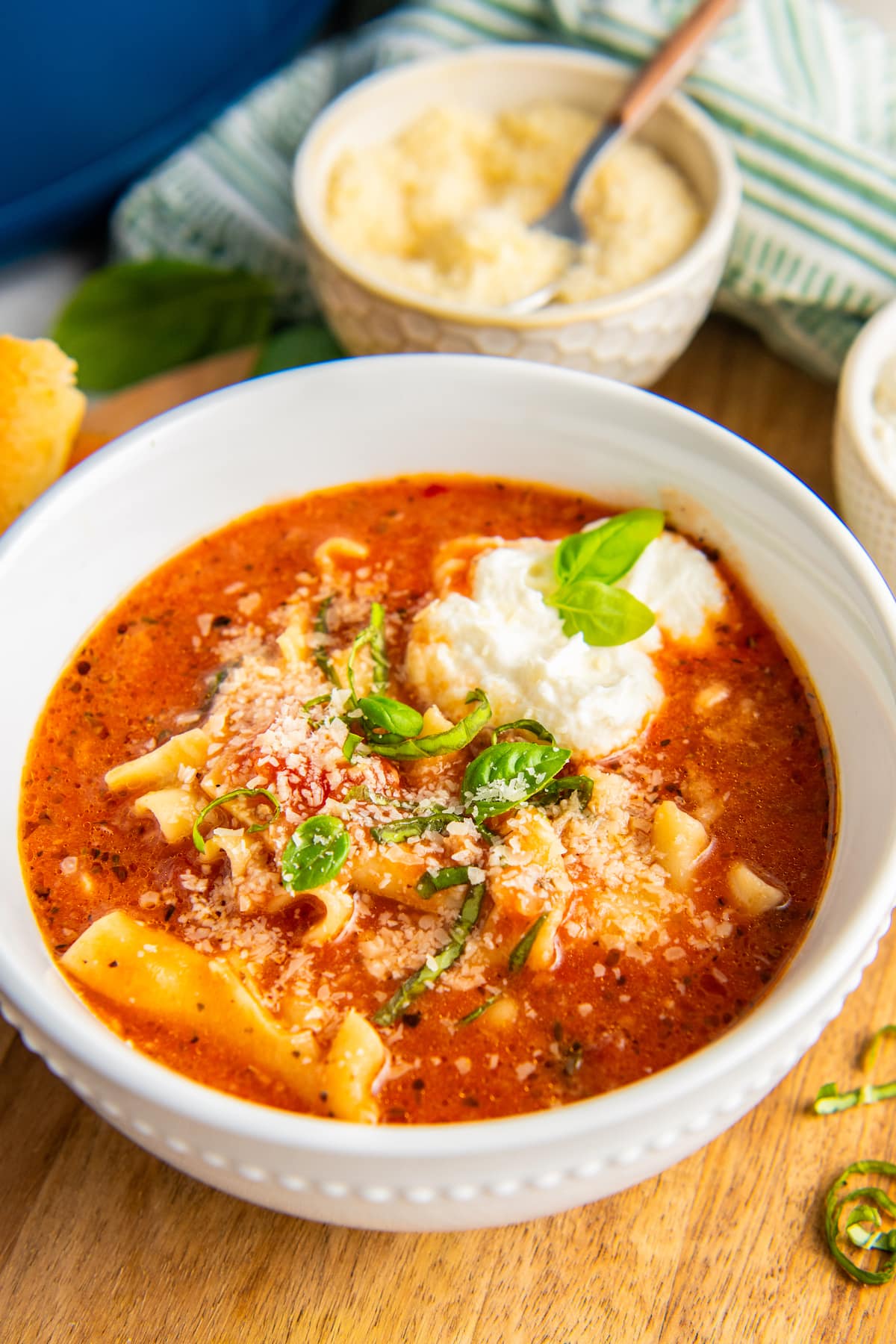 A bowl of lasagna soup topped with parmesan, ricotta, and basil, with a bowl of parmesan in the background