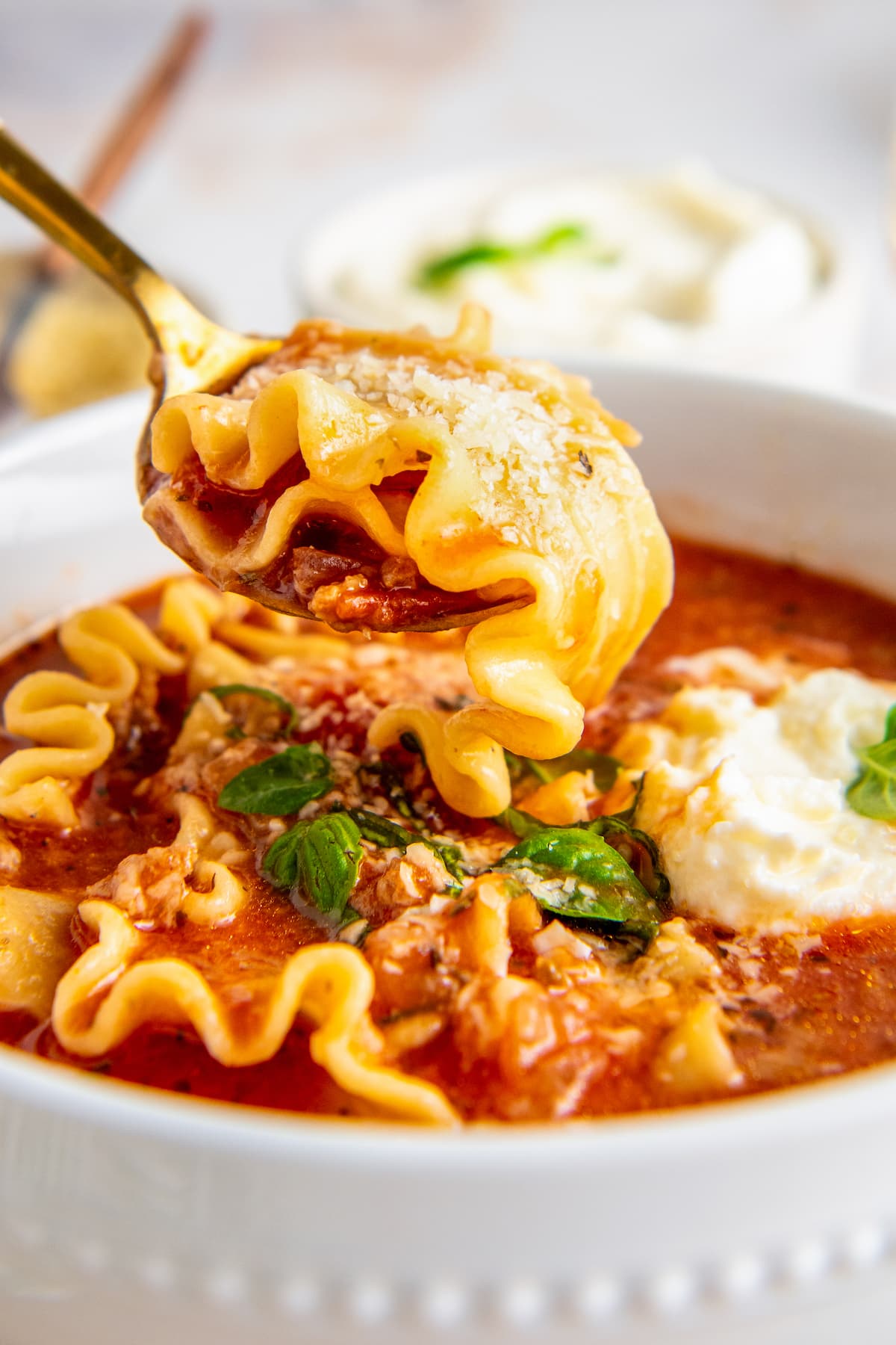 A spoon with a lasagna noodle in it, above a bowl of lasagna soup