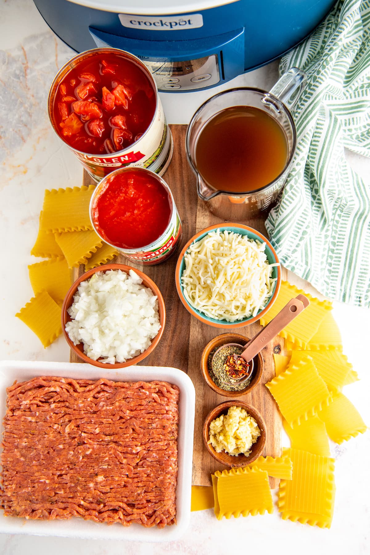 Overhead view of the ingredients for crockpot lasagna soup: Italian sausage, onions, diced tomatoes, tomato sauce, beef broth, mozzarella cheese, garlic, Italian seasoning, and broken up lasagna noodles, next to a crockpot