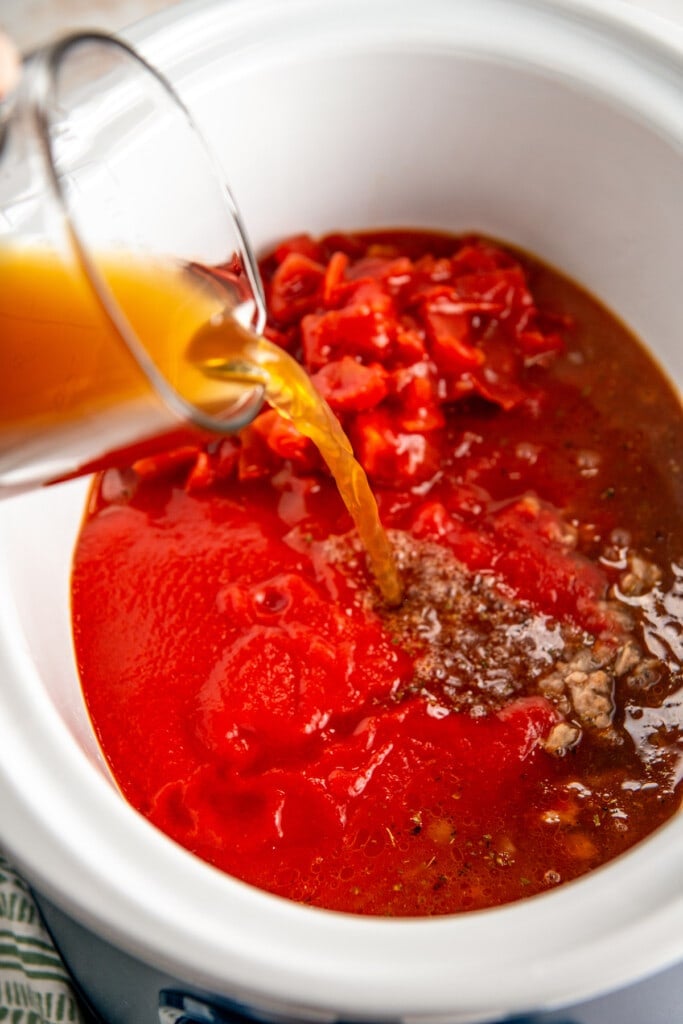 Beef broth being poured into a crockpot with diced tomatoes and tomato sauce
