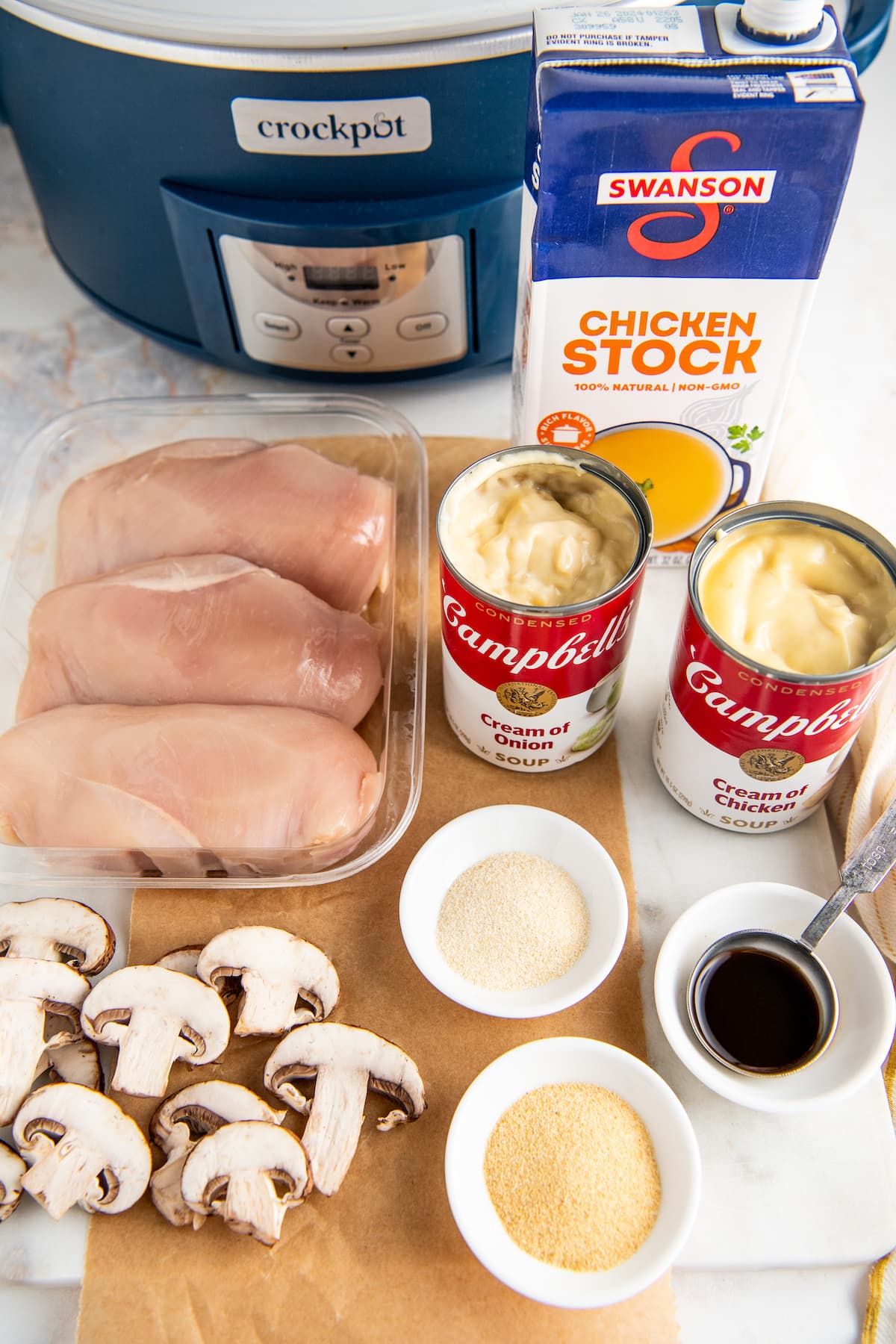 The ingredients needed for Crockpot Chicken & Gravy: sliced mushrooms, raw chicken breasts, a can of cream of chicken soup, a can of cream of onion soup, a container of chicken broth, a bowl of Worcestershire sauce, a bowl of garlic powder, and a bowl of onion powder, next to a crockpot.