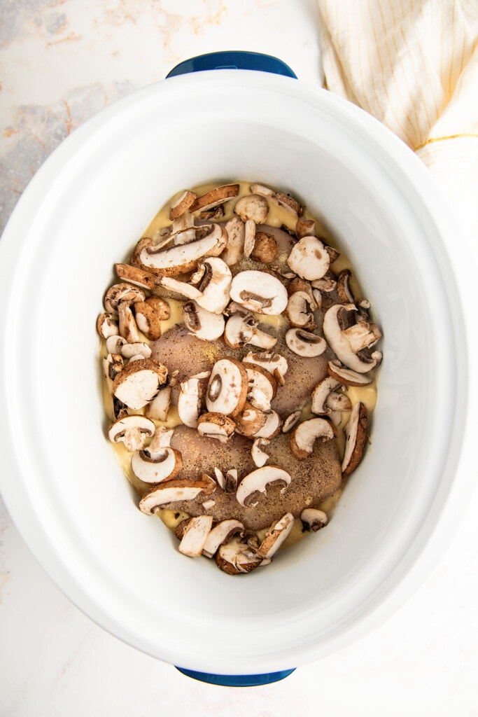 Overhead view of sliced raw mushrooms on top of raw chicken breasts in a crockpot.