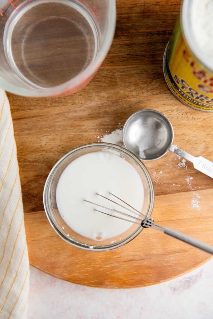 Overhead view of a jar with a slurry of cornstarch and water, with a whisk in it, next to a glass of water and a measuring spoon.