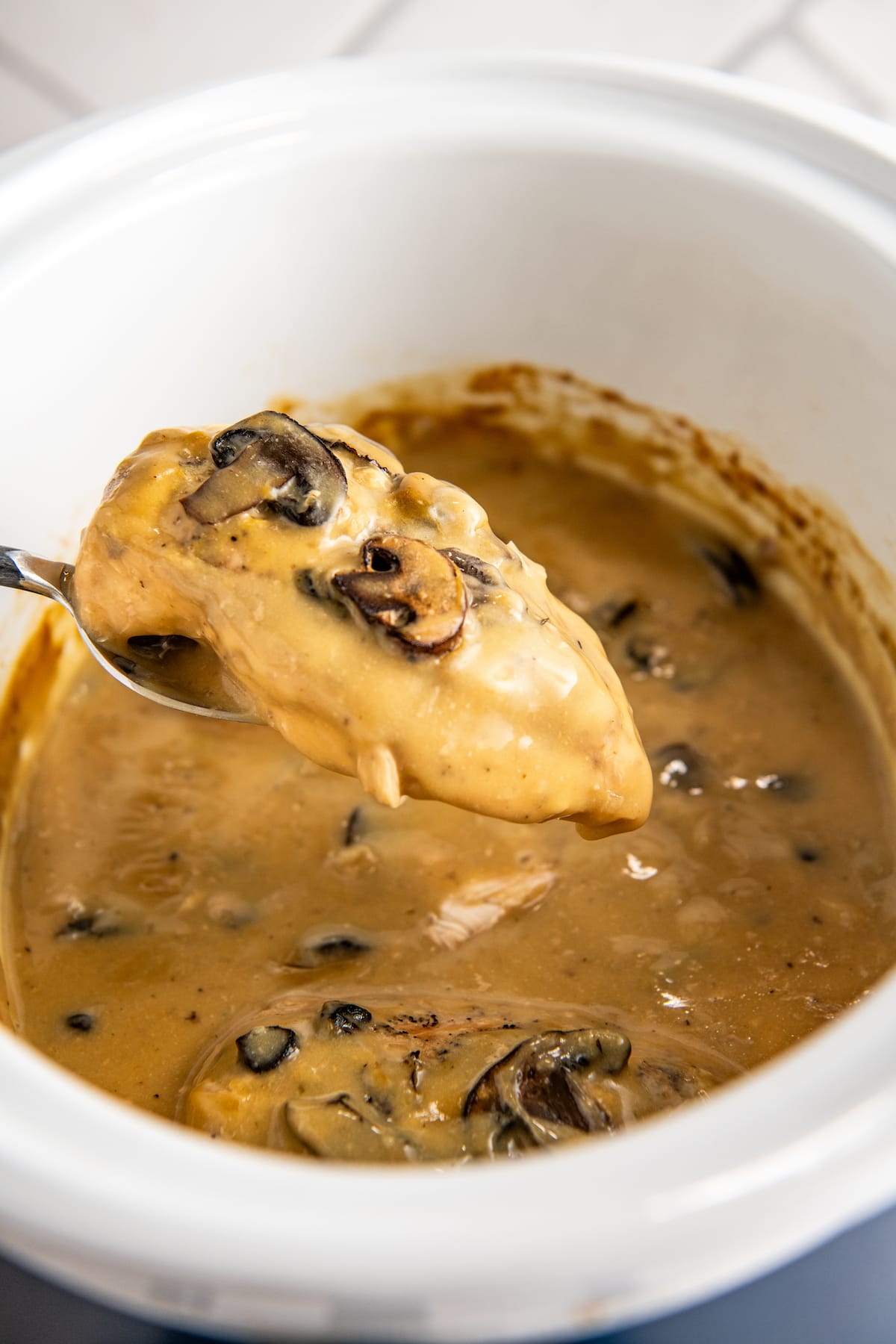 A spoon removing a chicken breast from a crockpot full of chicken and gravy with mushrooms.