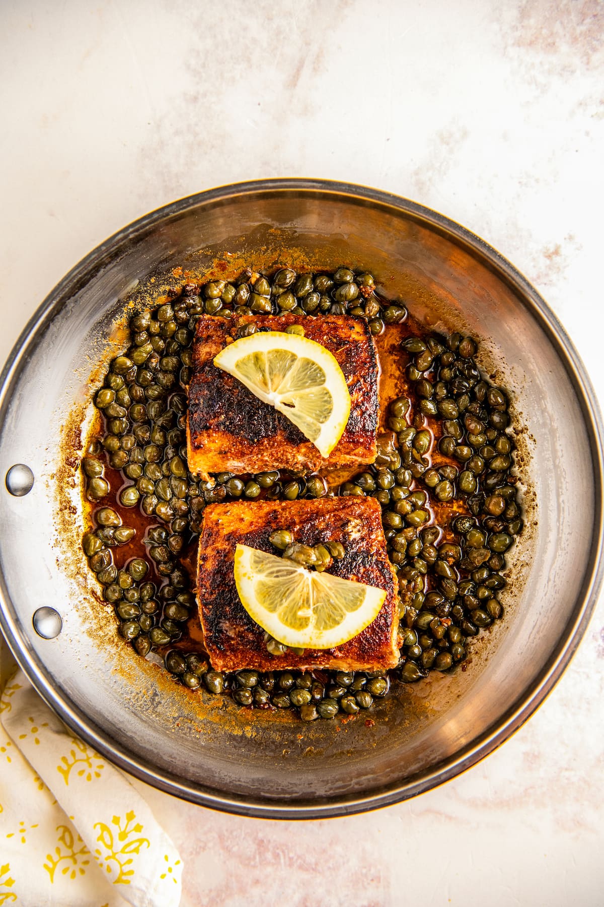 Overhead view of two mahi mahi fillets in a skillet on top of capers, topped with lemons