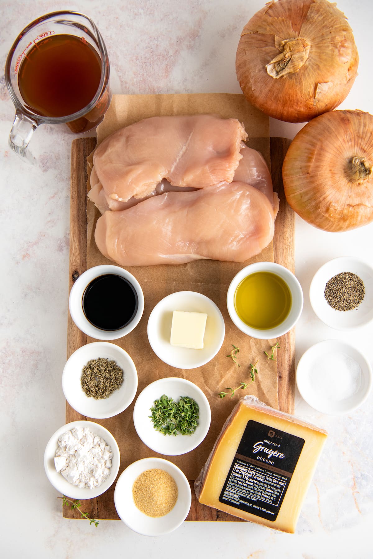 Overhead view of the ingredients needed for French onion chicken: chicken breasts, onions, a bowl of oil, a bowl of butter, a bowl of balsamic vinegar, a bowl of flour, a bowl of garlic powder, a bowl of pepper, a block of gruyere, a bowl of salt, a bowl of fresh thyme, a bowl of dried sage, and a pyrex of beef broth