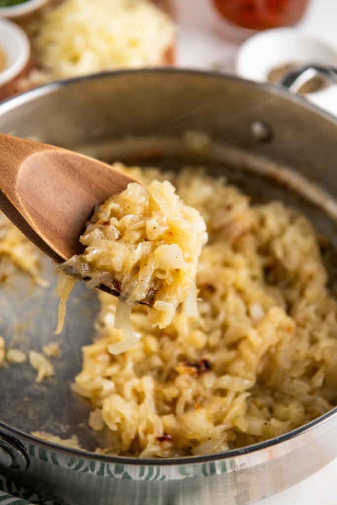 A wooden spoon full of caramelized onions, with a skillet full of them in the back.