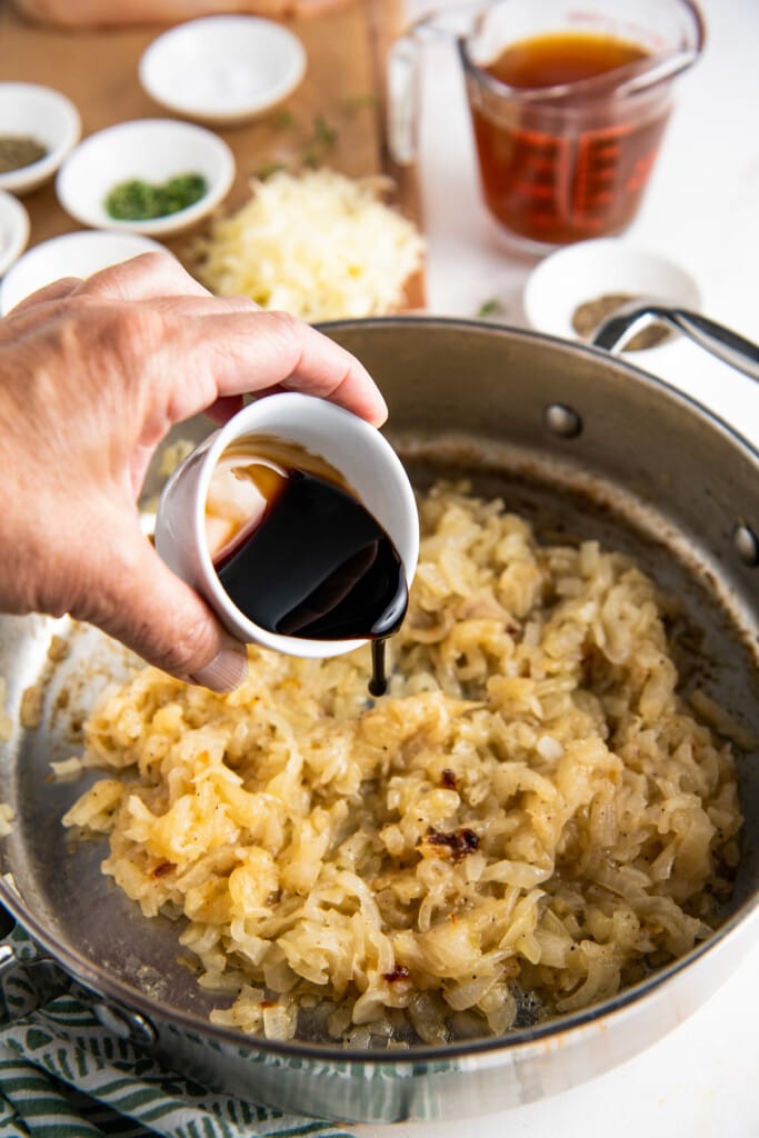 A hand pouring a bowl of balsamic vinegar into a skillet of caramelized onions
