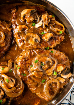 Overhead view of two steaks covered in gravy and mushrooms, cooking in a skillet