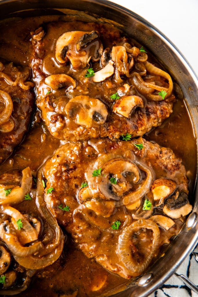 Overhead view of two steaks covered in gravy and mushrooms, cooking in a skillet