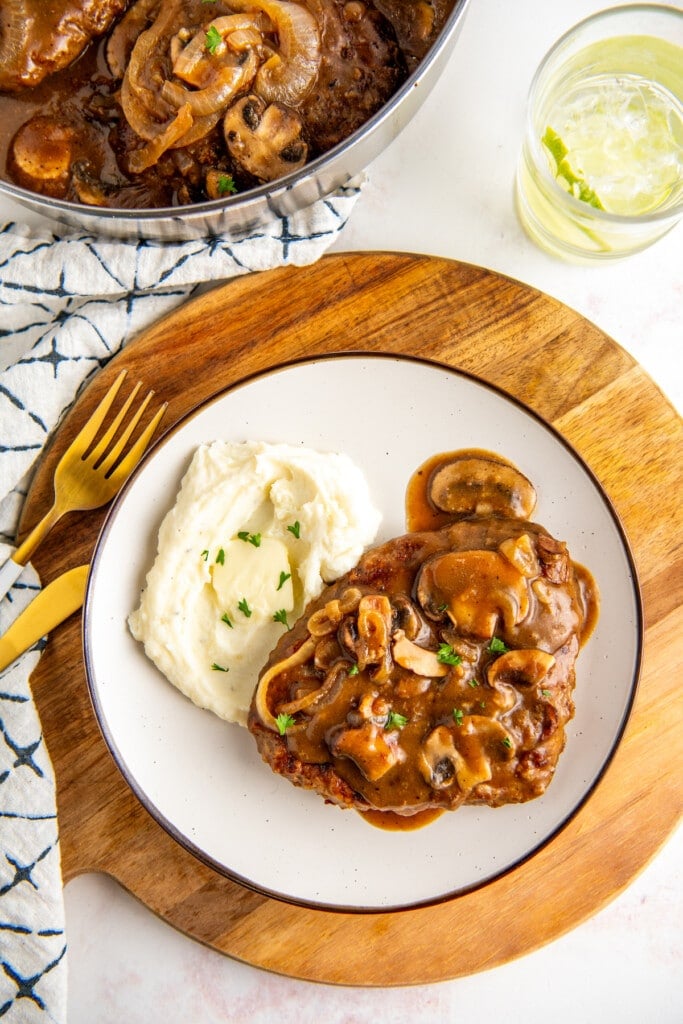 Overhead view of a plate with a steak smothered in mushrooms and gravy, and a pile of mashed potatoes, next to a fork.