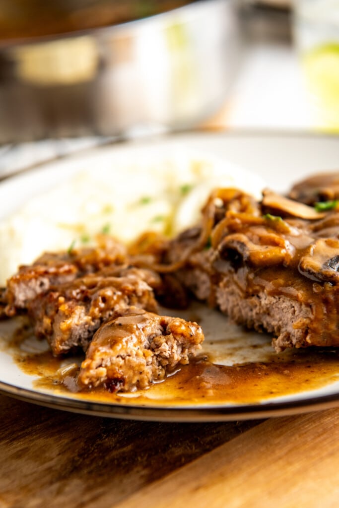 A smothered steak with mushrooms on a plate, with a few bites cut out.