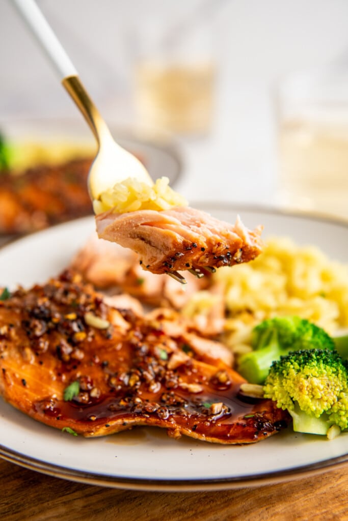 A piece of salmon on a fork, with a plate in the background full of salmon, broccoli, and rice