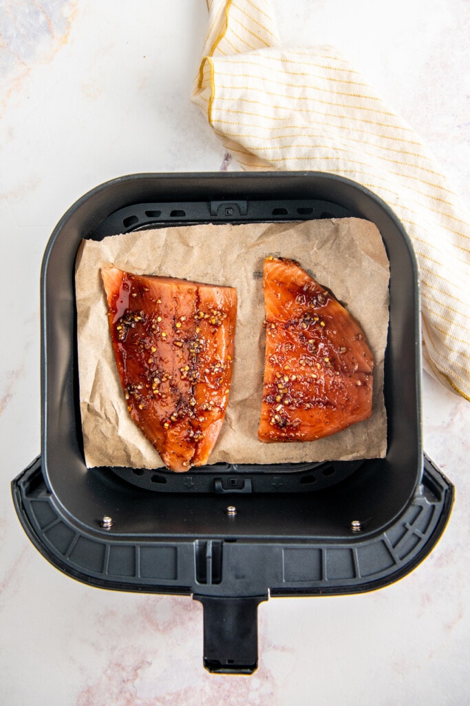 Two seasoned, raw salmon fillets on top of parchment paper in an air fryer basket