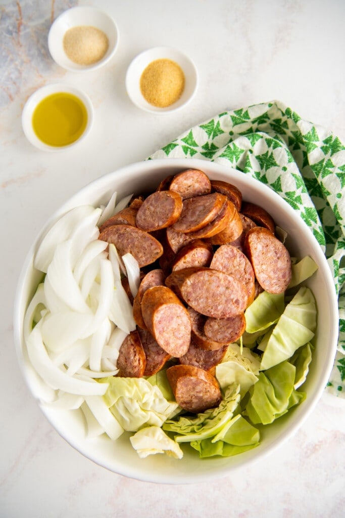 A mixing bowl filled with rounds of sausage, sliced cabbage, and sliced onions, next to a bowl of oil, a bowl of garlic powder, and a bowl of onion powder.