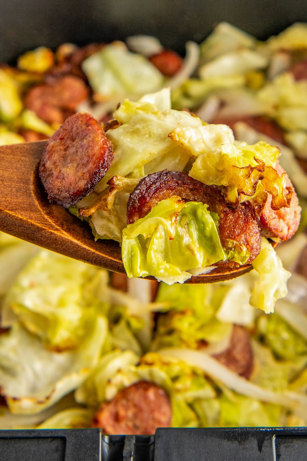 Close up of fried cabbage and sausage on a wooden spoon, above a an air fryer basket. 