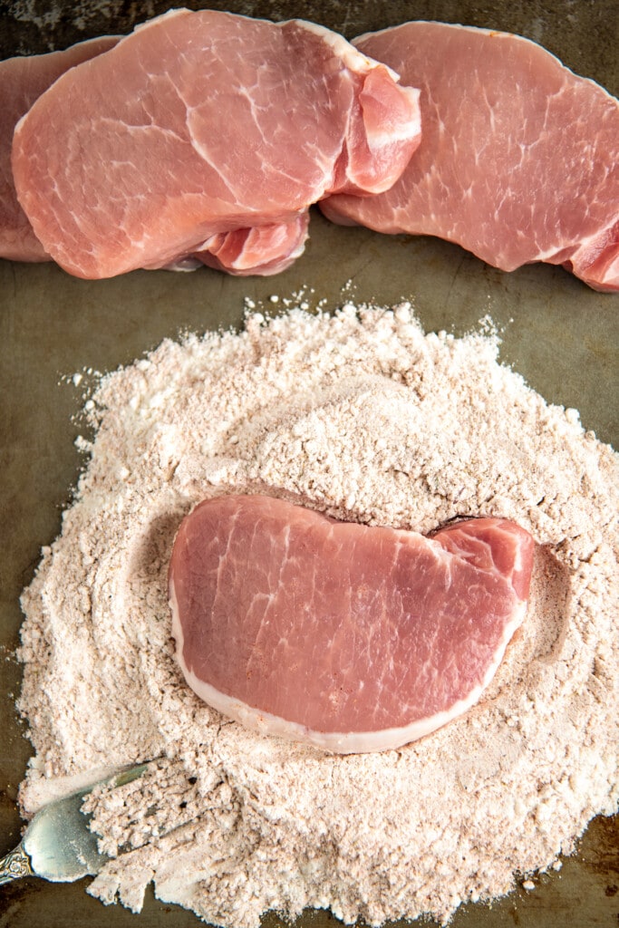 A raw pork chop in a pile of seasoned flour next to three more chops and a fork