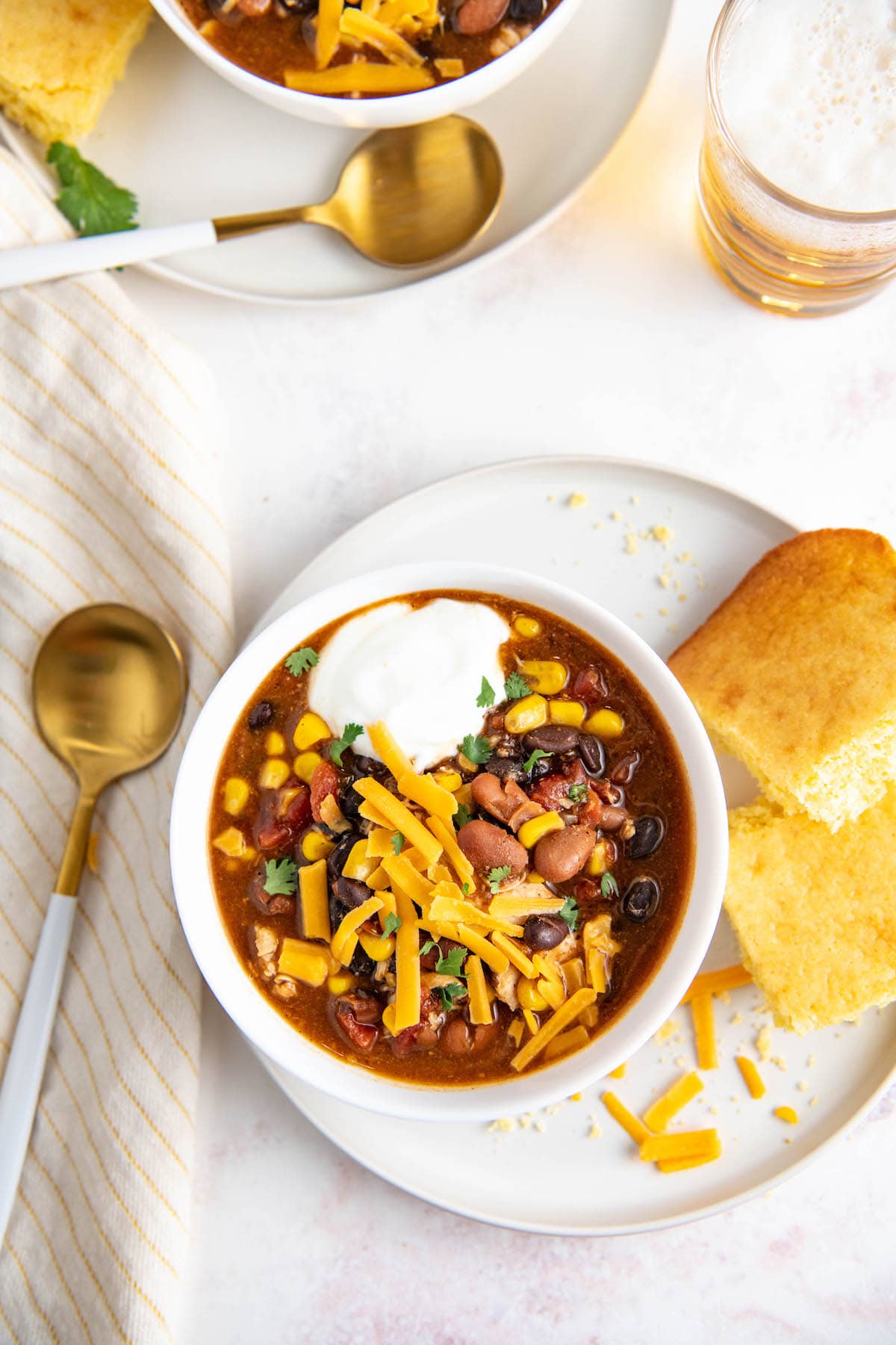 Overhead view of a bowl of chicken chili topped with cheese and sour cream on a bowl with corn bread.