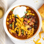 Overhead view of a bowl of chicken chili topped with cheese and sour cream, on a plate with cornbread