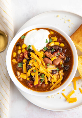 Overhead view of a bowl of chicken chili topped with cheese and sour cream, on a plate with cornbread