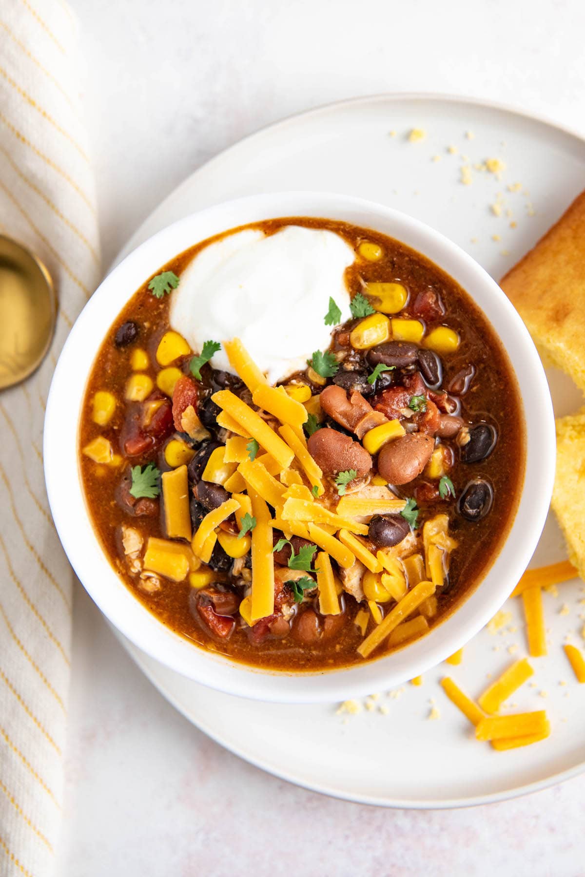 Overhead view of a bowl of chicken chili topped with cheese and sour cream, on a plate with cornbread.