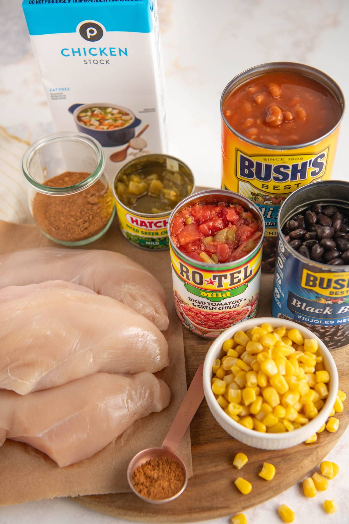 The ingredients needed for crockpot chicken chili: raw chicken breasts, a bowl of corn, a can of black beans, a can of Rotel, a can of chili beans, a can of green chilis, a container of chicken broth, and a bowl of chili seasoning.