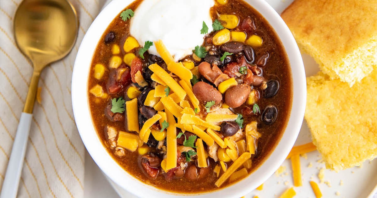 Close up of a bowl of chicken chili topped with cheese and sour cream, with a spoon and cornbread on the side