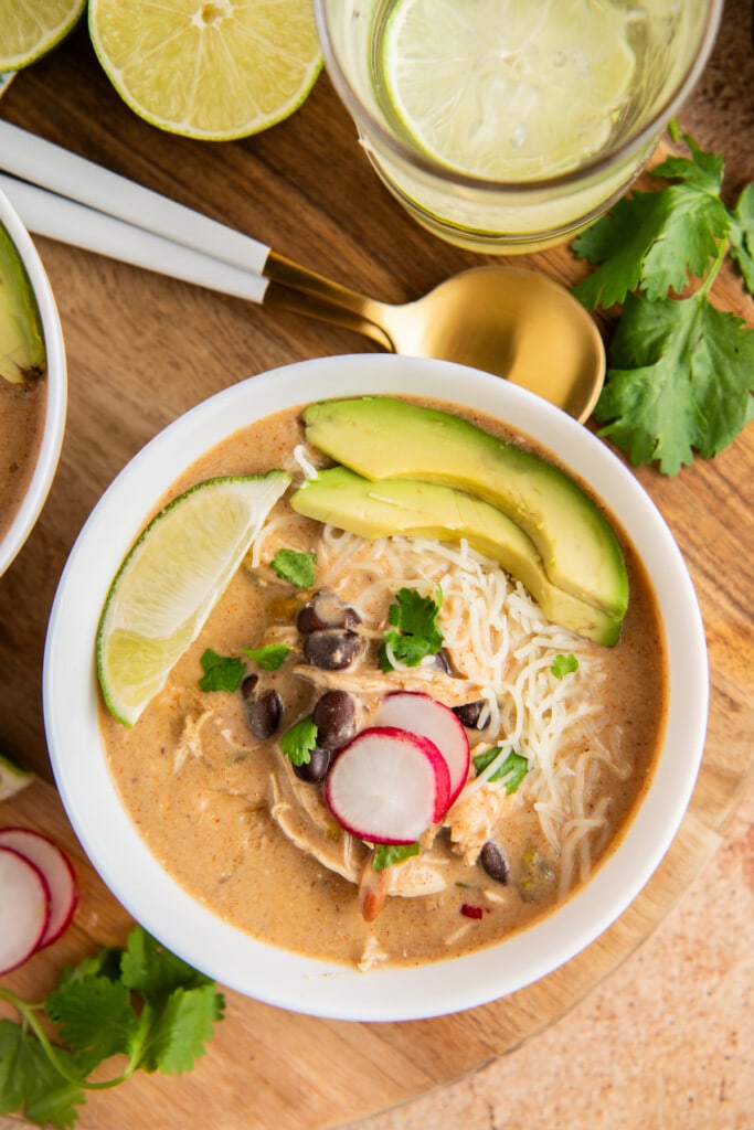 Overhead view of a bowl of slow cooker chicken enchilada soup topped with avocado, radish, cilantro, and lime, next to a spoon and cilantro.