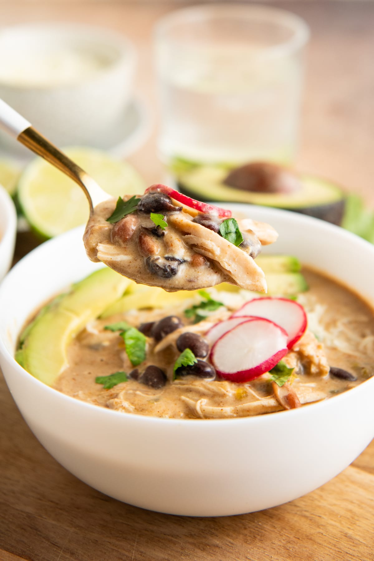 A spoon with a spoonful of soup in it, above a bowl of chicken enchilada soup topped with radishes and avocado, with a glass of water and half an avocado in the background.
