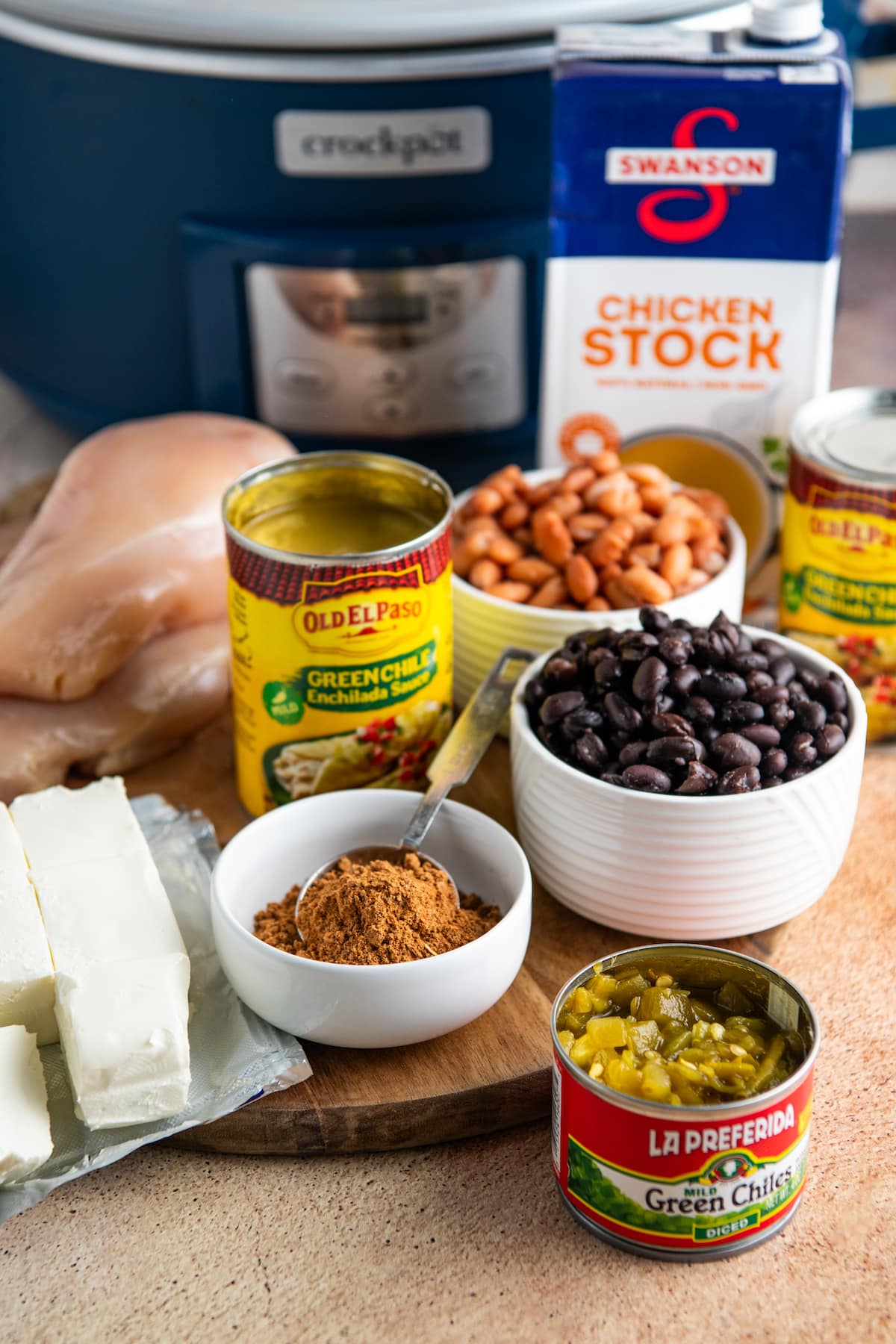 All the ingredients needed for chicken enchilada soup: a bowl of pinto beans, a bowl of black beans, a can of enchilada sauce, a can of green chiles, a bowl of taco seasoning, a carton of chicken stock, a block of cream cheese, and raw chicken breasts, in front of a crockpot.