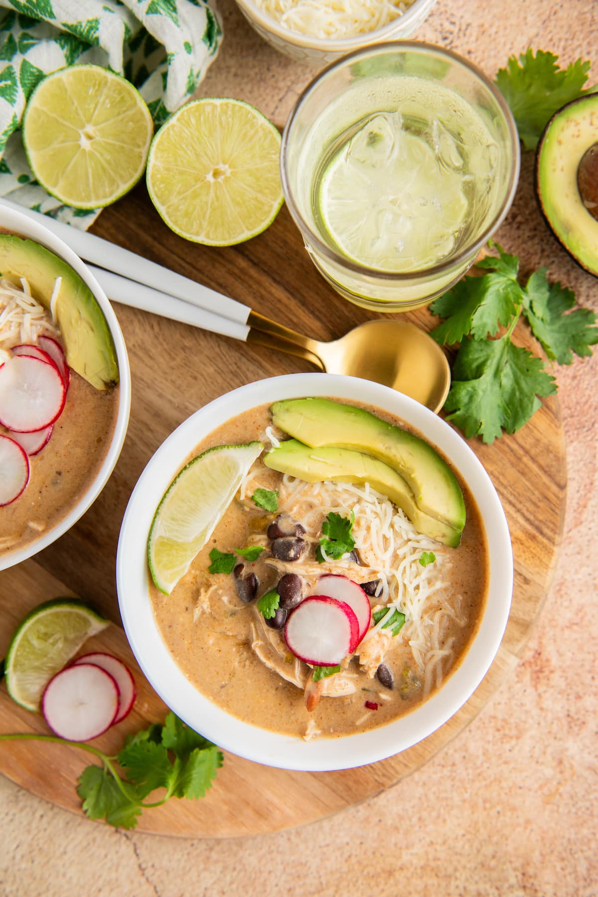 Overhead view of a bowl of crock pot chicken enchilada soup topped with avocado, radish, lime, and cilantro, next to another bowl, a lime cut in half, sliced radishes, fresh cilantro, and a glass of water.
