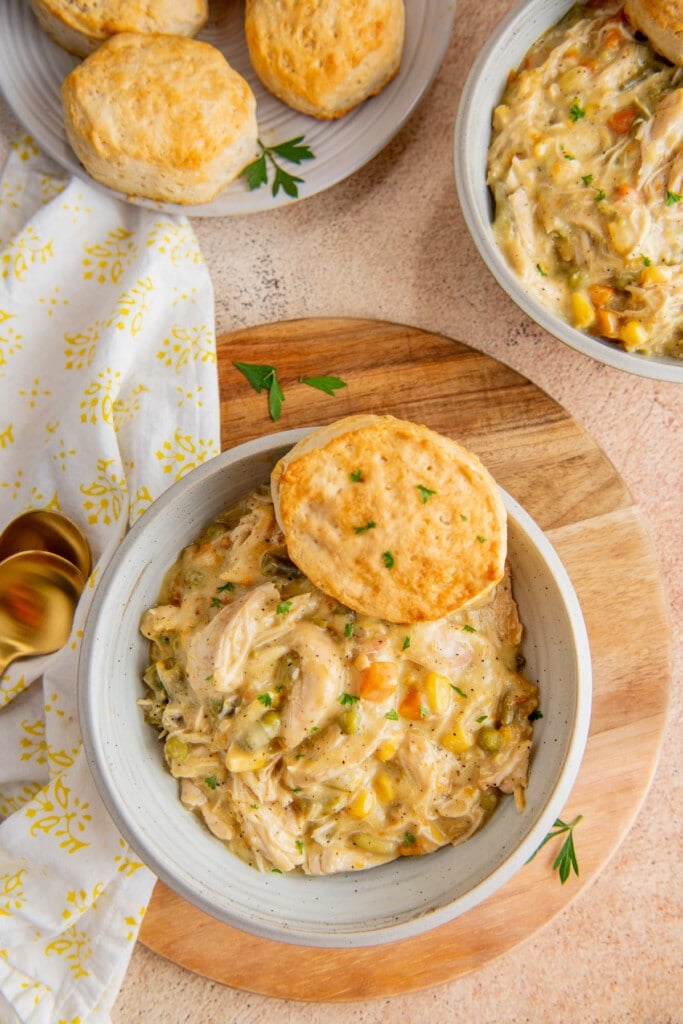 Overhead view of a bowl of chicken pot pie filling with a biscuit, on a cutting board, next to another bowl of it and a plate of biscuits