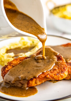 A gravy boat pouring gravy onto a pork chop with mashed potatoes in the backrgound