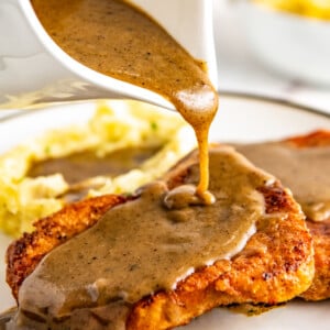 A gravy boat pouring gravy onto a pork chop with mashed potatoes in the backrgound