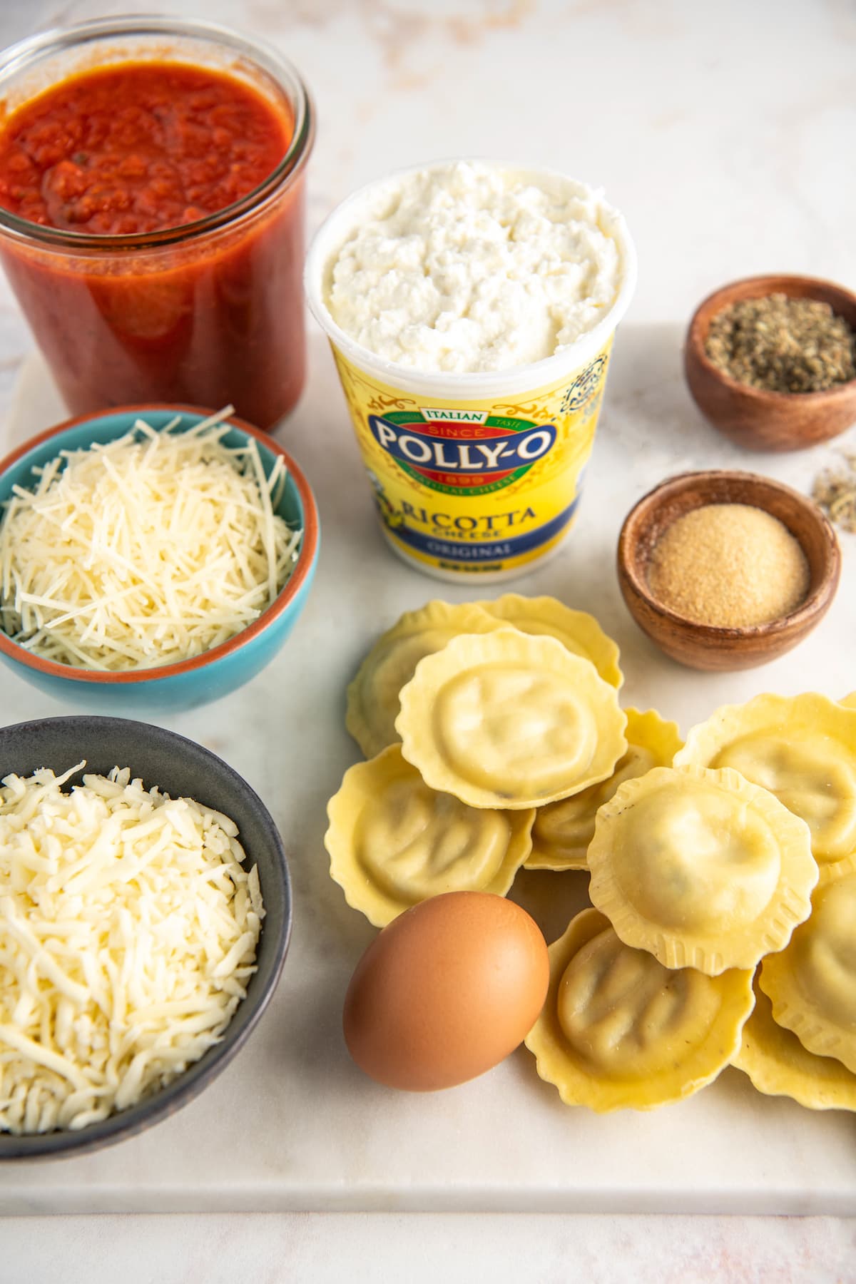 All the ingredients needed for ravioli lasagna: a pile of fresh ravioli, an egg, a carton of ricotta, a bowl of shredded parmesan, a bowl of shredded mozzarella, a jar of tomato sauce, a bowl of garlic powder, and a bowl of Italian seasoning