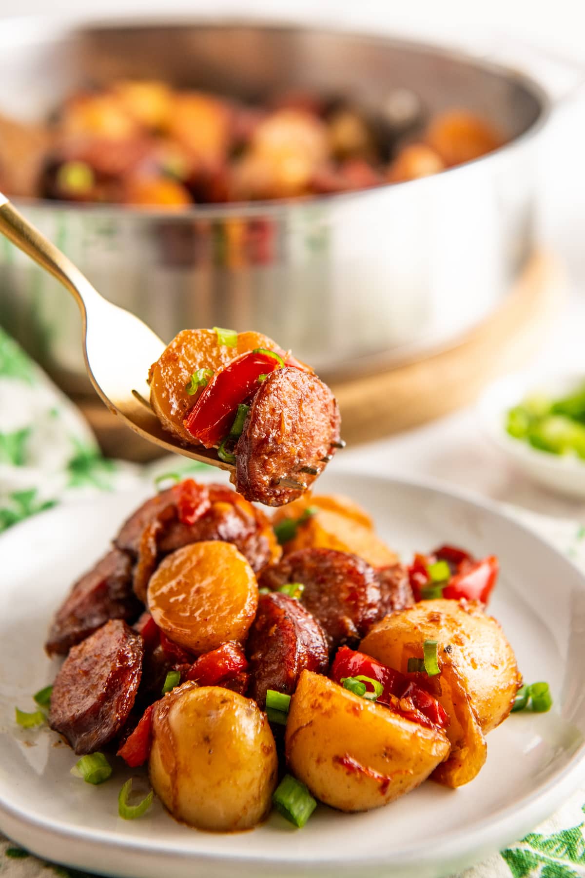 A plate of sausage and potatoes with a fork lifting up a bite, with a serving bowl in the background