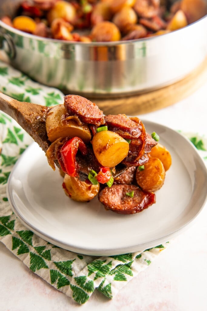 A wooden spoon putting a serving of sausage and potatoes on a plate, with a serving bowl in the background